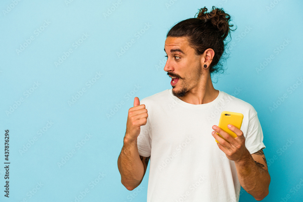 Young caucasian man with long hair holding mobile phone isolated on blue background points with thumb finger away, laughing and carefree.