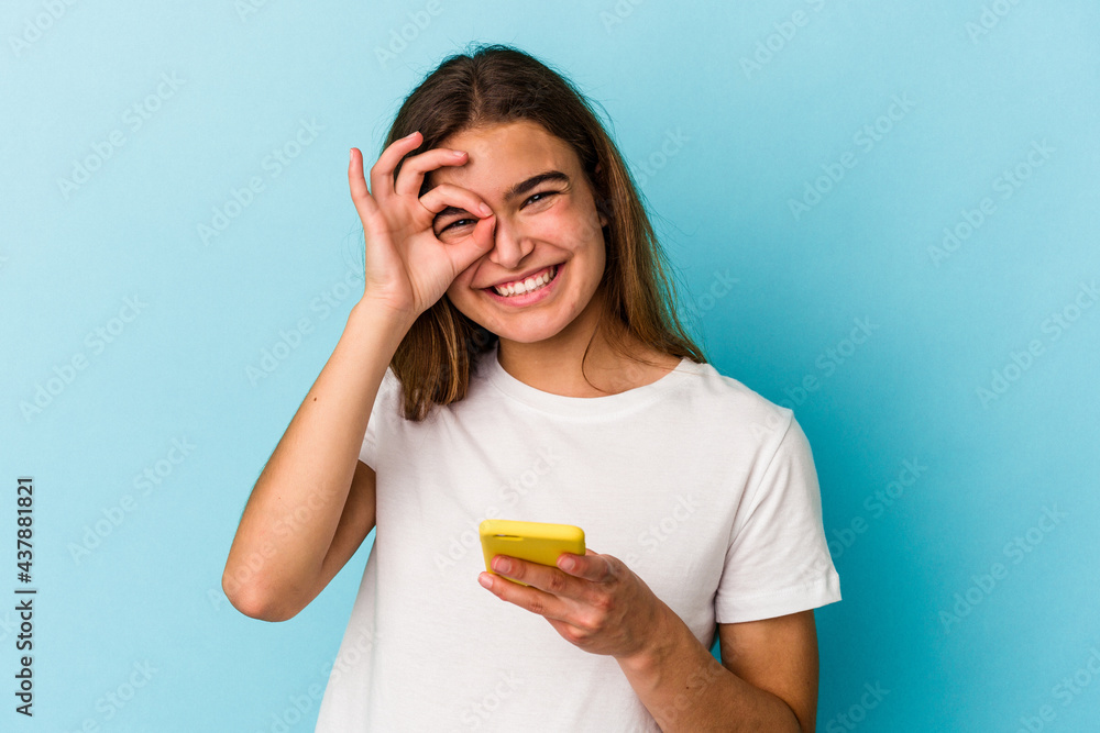 Young caucasian woman holding a mobile phone isolated on blue background excited keeping ok gesture on eye. Young caucasian woman holding a mobile phone isolated on blue background excited keeping ok gesture on eye.