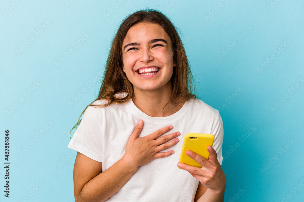 Young caucasian woman holding a mobile phone isolated on blue background laughs out loudly keeping hand on chest. Young caucasian woman holding a mobile phone isolated on blue background laughs out loudly keeping hand on chest.