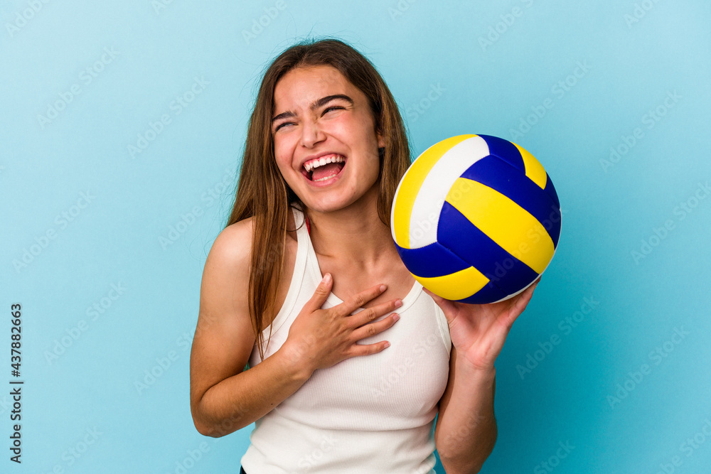Young caucasian woman playing volleyball isolated on blue background laughs out loudly keeping hand on chest.