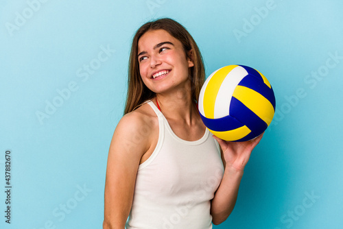 Young caucasian woman playing volleyball isolated on blue background looks aside smiling, cheerful and pleasant.