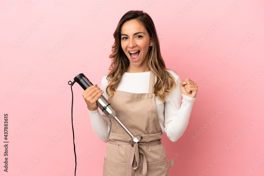 Young woman using hand blender over isolated pink background celebrating a victory in winner position Young woman using hand blender over isolated pink background celebrating a victory in winner position