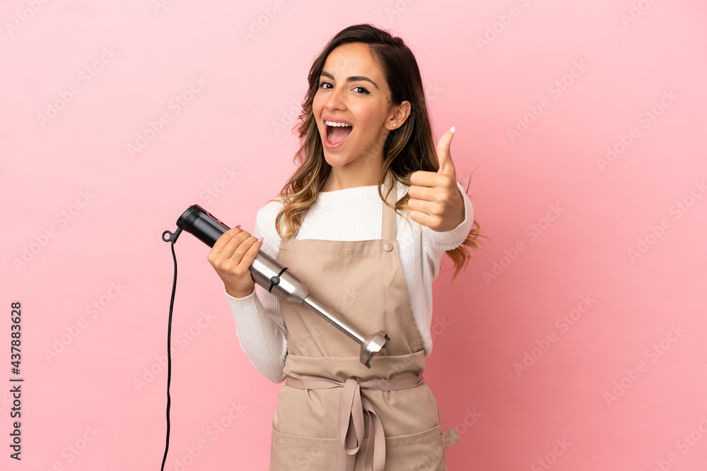 Young woman using hand blender over isolated pink background with thumbs up because something good has happened Young woman using hand blender over isolated pink background with thumbs up because something good has happened