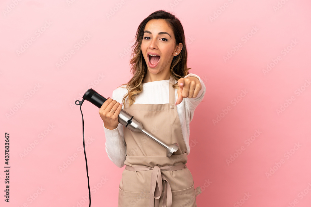 Young woman using hand blender over isolated pink background surprised and pointing front Young woman using hand blender over isolated pink background surprised and pointing front