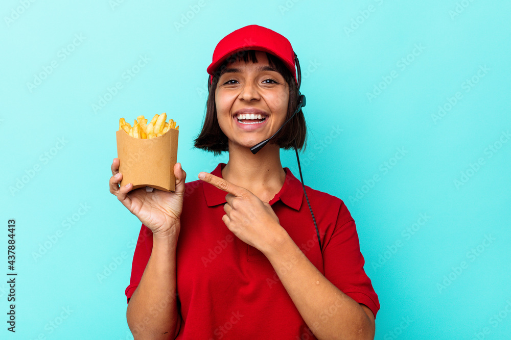 Young mixed race woman fast food restaurant worker holding fries isolated on blue background smiling and pointing aside, showing something at blank space. Young mixed race woman fast food restaurant worker holding fries isolated on blue background smiling and pointing aside, showing something at blank space.