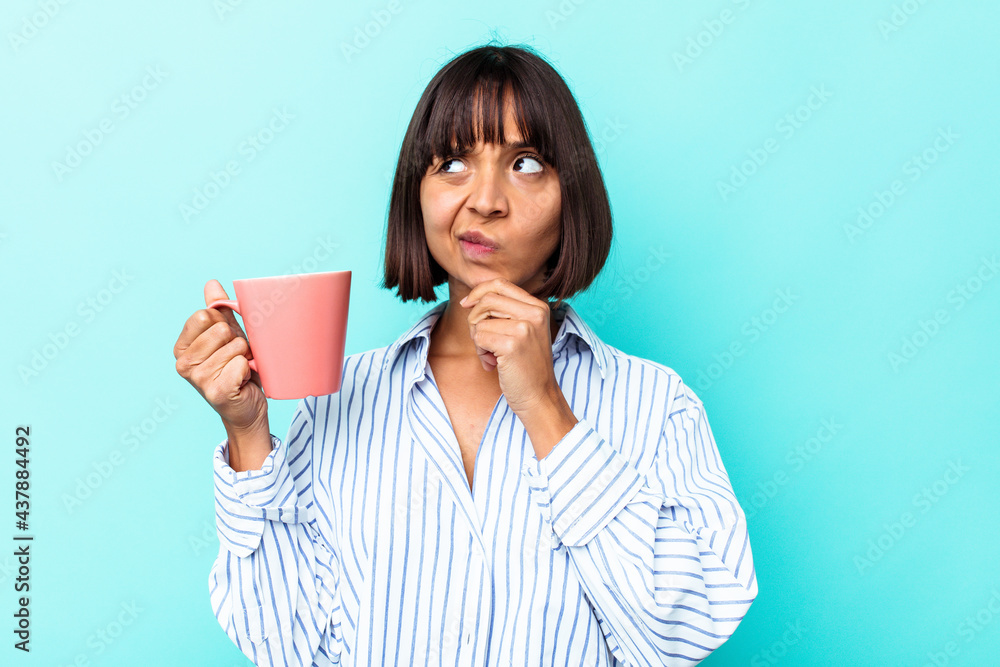 Young mixed race woman holding a pink mug isolated on blue background looking sideways with doubtful and skeptical expression. Young mixed race woman holding a pink mug isolated on blue background looking sideways with doubtful and skeptical expression.
