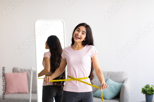 Indian woman measuring her waist with tape in front of mirror, showing results of slimming diet or liposuction at home photo