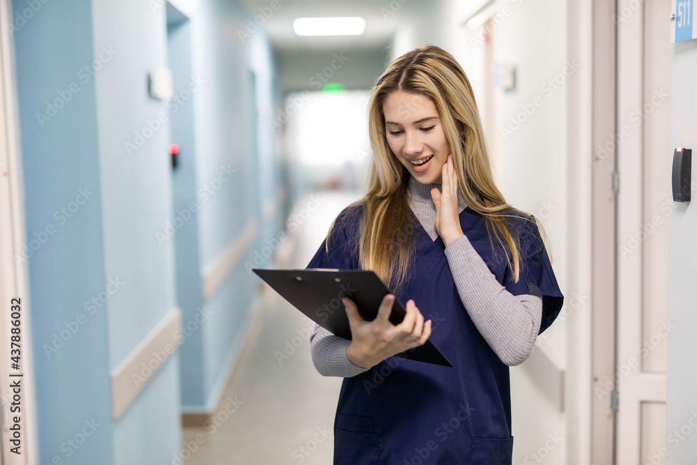 Portrait Of Smiling Female Doctor Wearing Scrubs Standing In Hospital Corridor Holding Clipboard Portrait Of Smiling Female Doctor Wearing Scrubs Standing In Hospital Corridor Holding Clipboard