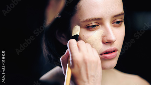 makeup artist holding cosmetic brush while applying makeup foundation on skin of young model