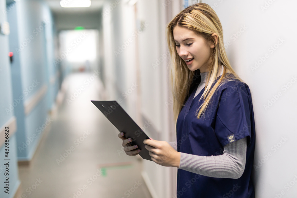 Portrait Of Smiling Female Doctor Wearing Scrubs Standing In Hospital Corridor Holding Clipboard Portrait Of Smiling Female Doctor Wearing Scrubs Standing In Hospital Corridor Holding Clipboard