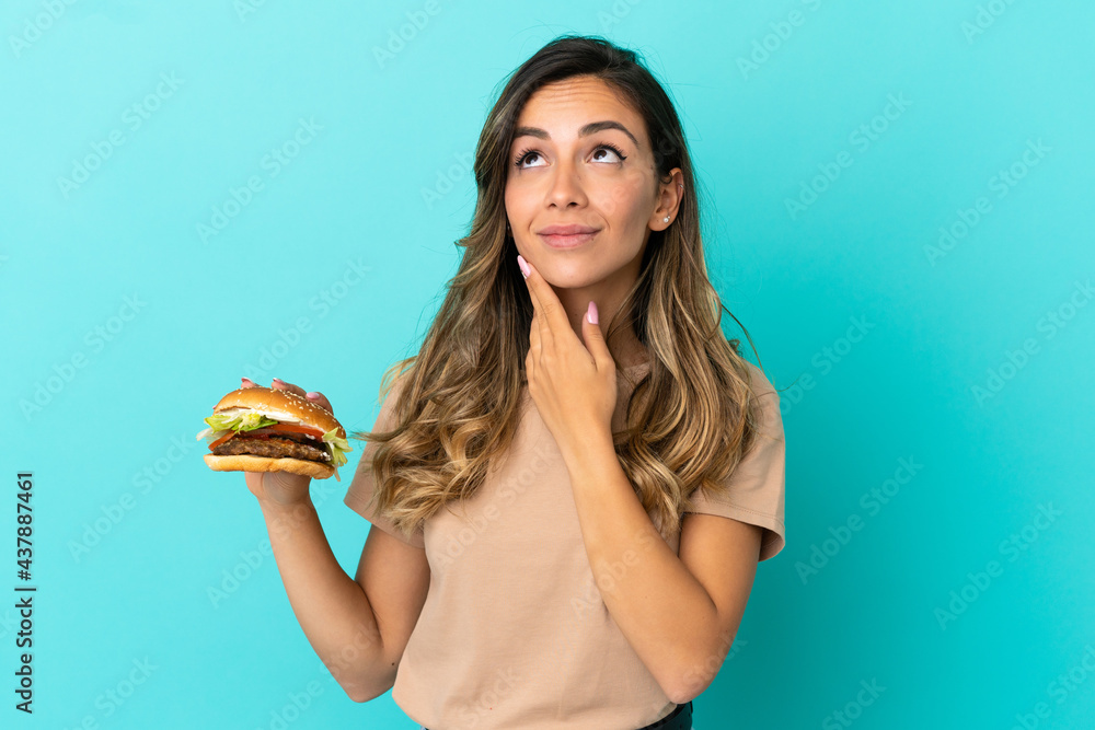 Young woman holding a burger over isolated background looking up while smiling Young woman holding a burger over isolated background looking up while smiling