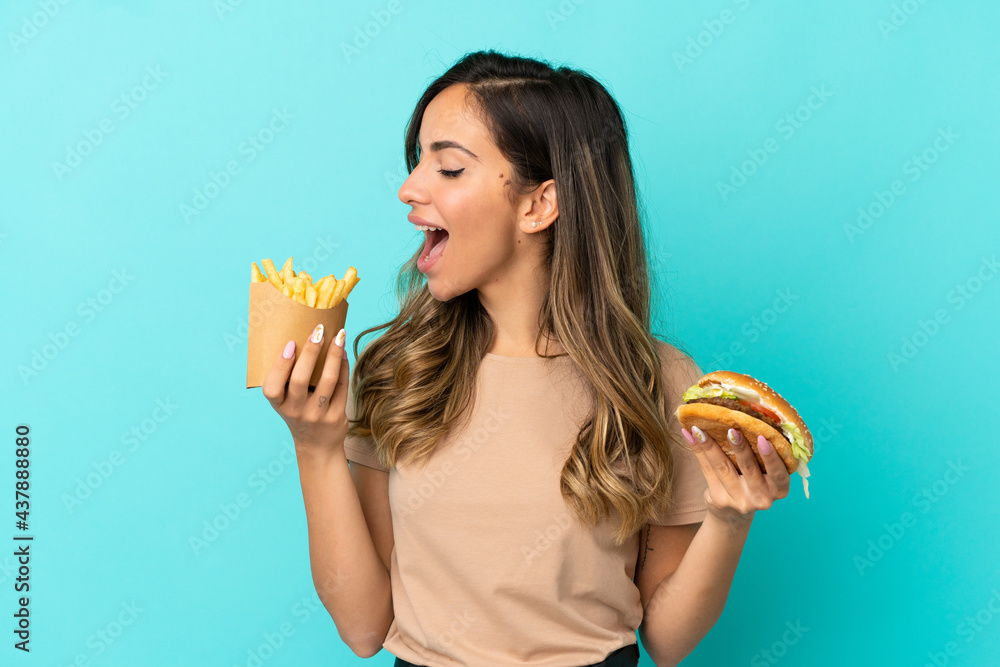 Young woman holding fried chips and burger over isolated background Young woman holding fried chips and burger over isolated background