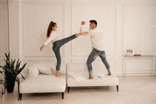 Fotografie young couple in safety masks fighting on couch while fooling around at home