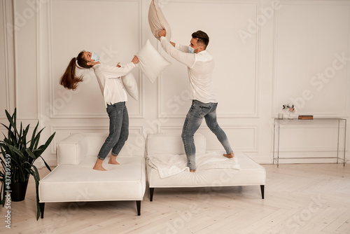 young couple in medical masks fighting with pillows while having fun at home