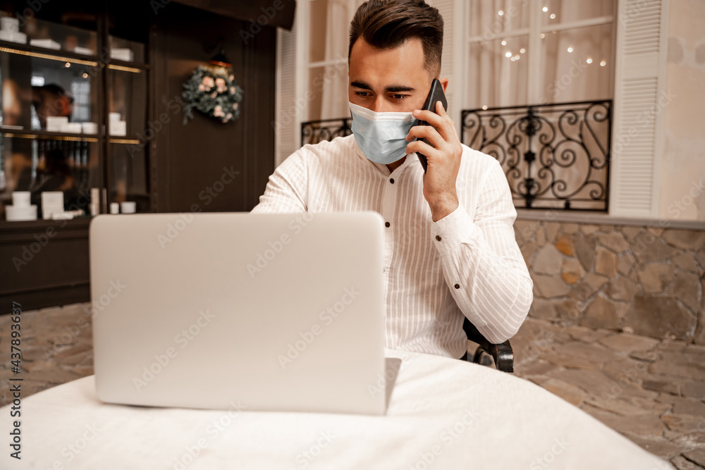 young man in medical mask working on laptop while talking on smartphone in cafe young man in medical mask working on laptop while talking on smartphone in cafe