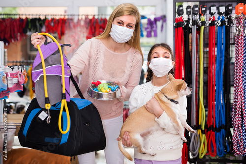 Child girl with her mother in face masks in pet shop looking through different clothes and accessories for their dog