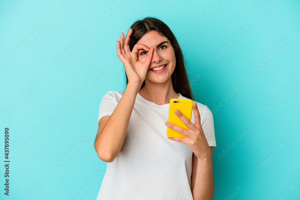 Young caucasian woman holding a mobile phone isolated on blue background excited keeping ok gesture on eye. Young caucasian woman holding a mobile phone isolated on blue background excited keeping ok gesture on eye.