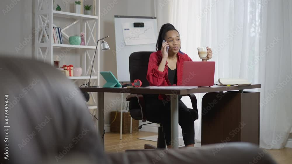 Wide shot portrait of positive African American young woman drinking morning cappuccino talking on smartphone sitting at table in home office. Smiling beautiful millennial planning work time indoors Wide shot portrait of positive African American young woman drinking morning cappuccino talking on smartphone sitting at table in home office. Smiling beautiful millennial planning work time indoors