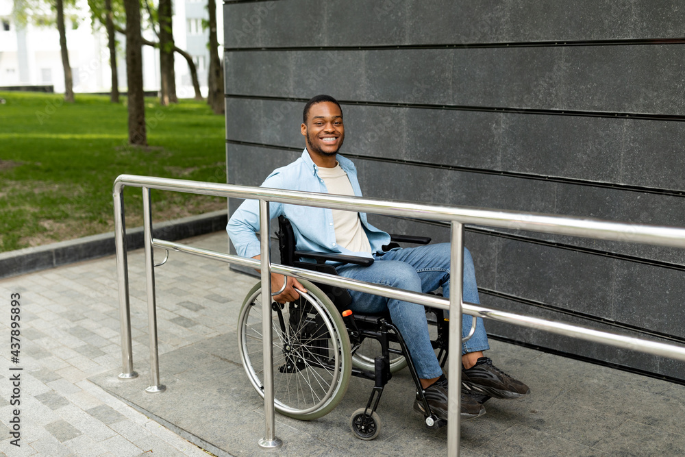 Obraz premium Happy impaired black man in wheelchair entering building on ramp outdoors, full length. Handicapped-accessible city