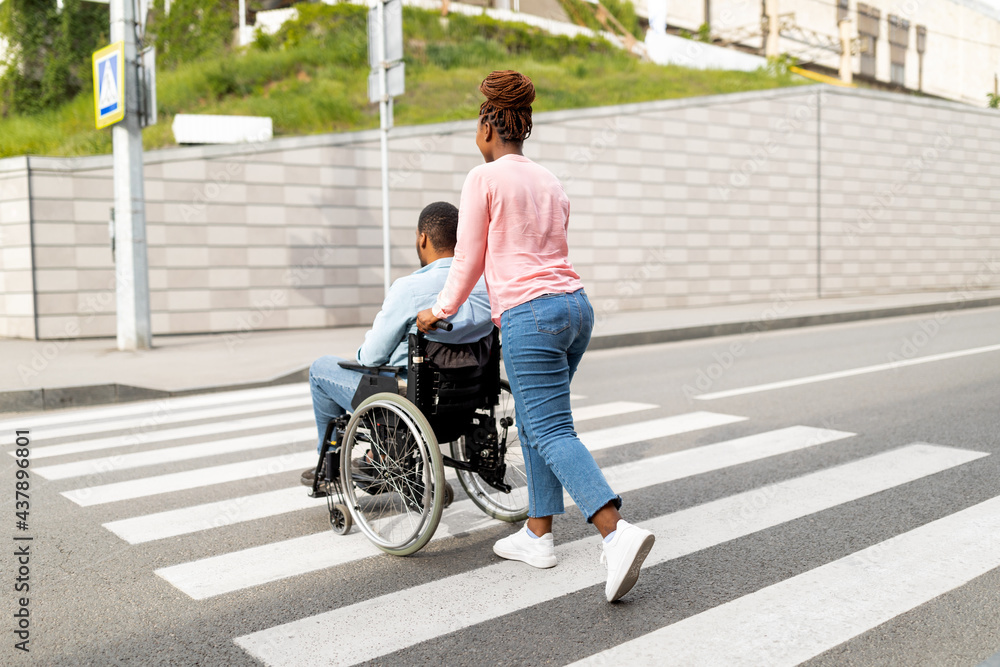 Back view of young female caregiver pushing wheelchair with black male patient across city street, full length