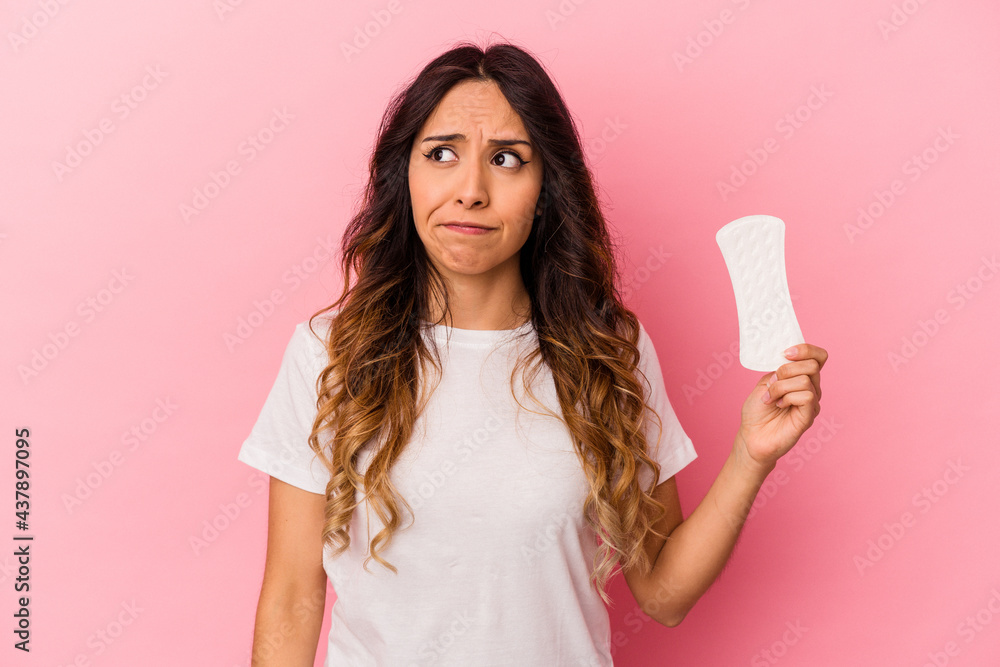 Young mexican woman holding a compress isolated on pink background confused, feels doubtful and unsure. Young mexican woman holding a compress isolated on pink background confused, feels doubtful and unsure.