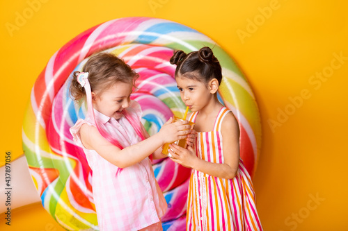 two happy little girls in colorful dress drinking orange juice having fun on yellow background with lollipop