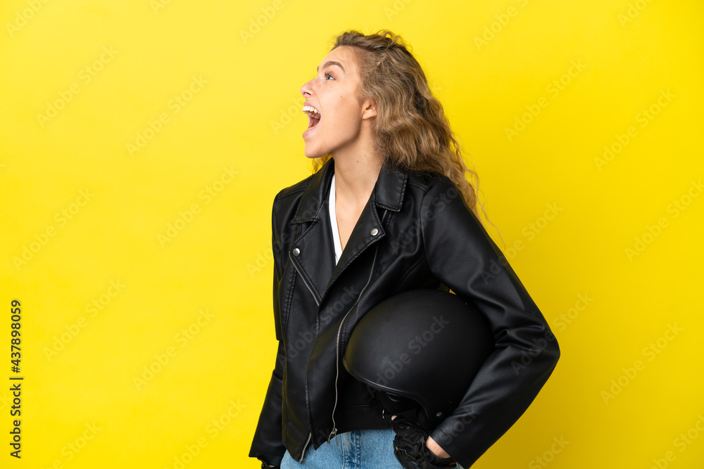 Young blonde woman with a motorcycle helmet isolated on yellow background laughing in lateral position Young blonde woman with a motorcycle helmet isolated on yellow background laughing in lateral position