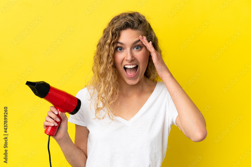 Young blonde woman holding a hairdryer isolated on yellow background with surprise expression Young blonde woman holding a hairdryer isolated on yellow background with surprise expression