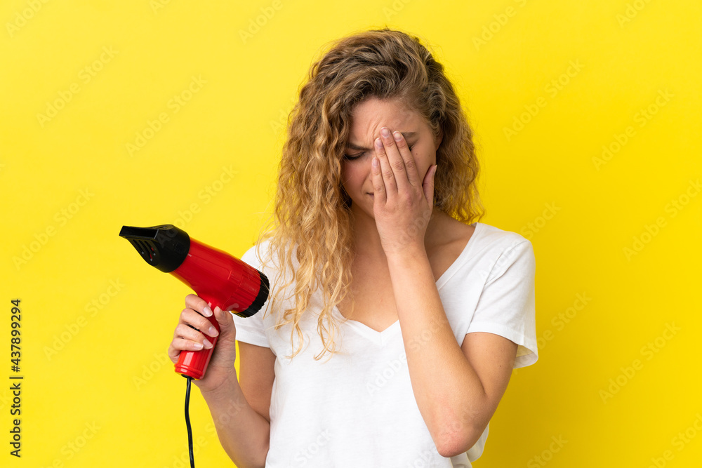 Young blonde woman holding a hairdryer isolated on yellow background with tired and sick expression Young blonde woman holding a hairdryer isolated on yellow background with tired and sick expression
