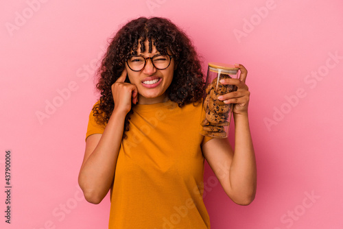 Young mixed race woman holding a cookies jar isolated on pink background covering ears with hands.