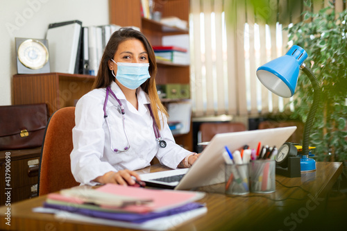 Woman doctor in protective mask sitting at workplace with computer in her office