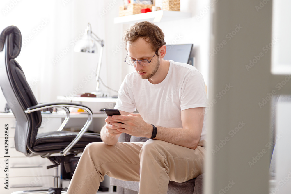 Happy young man in eyeglasses sitting on sofa sending text on mobile phone Happy young man in eyeglasses sitting on sofa sending text on mobile phone