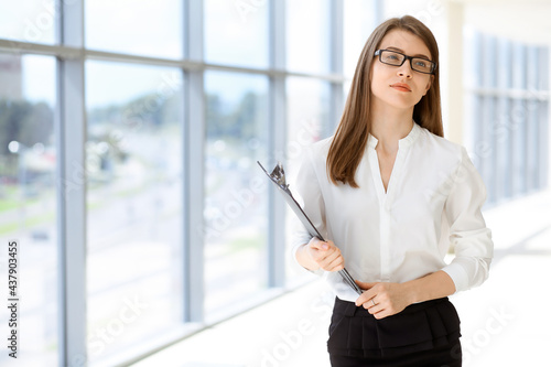 Beautiful female specialist with clipboard standing in modern office and smiling charmingly. Working on design, data analysis, plan strategy. Business people concept © volha_r