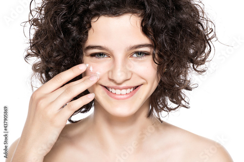 Teenager skincare. Beautiful teenage girl with gorgeous curly hair applying moisturiser face cream, looking at camera smiling. Studio shot on white background. Beauty, skincare and puberty concept.