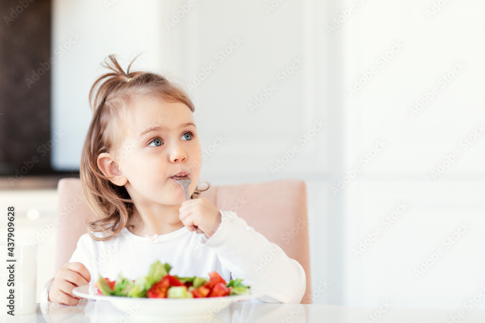 Child girl eating fresh raw vegetables vitamin salad in white kitchen background and thoughtfully looks at the empty copy space.Healthy nutrition food for children. Place for text for advertising. Child girl eating fresh raw vegetables vitamin salad in white kitchen background and thoughtfully looks at the empty copy space.Healthy nutrition food for children. Place for text for advertising.