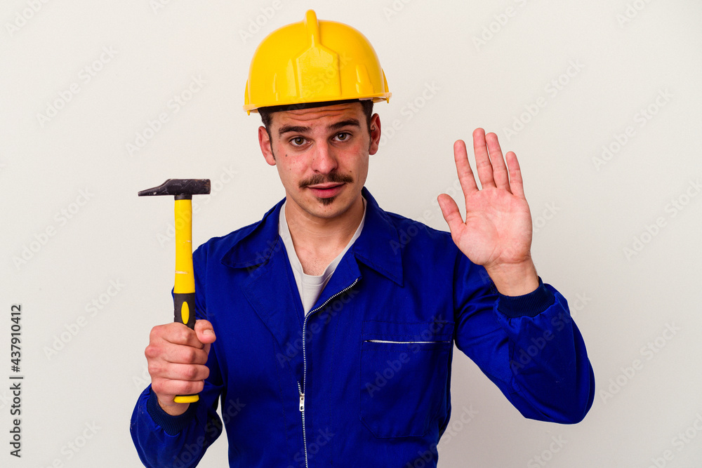 Young caucasian worker man holding a hammer isolated on white background smiling cheerful showing number five with fingers. Young caucasian worker man holding a hammer isolated on white background smiling cheerful showing number five with fingers.