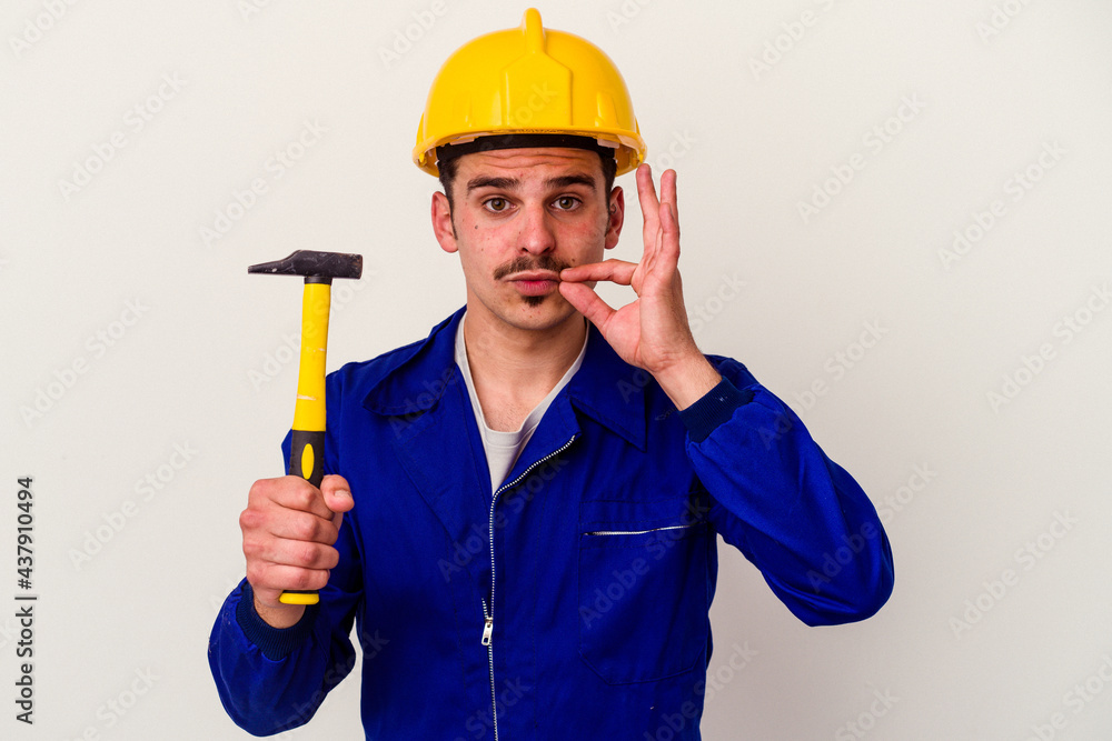 Young caucasian worker man holding a hammer isolated on white background with fingers on lips keeping a secret. Young caucasian worker man holding a hammer isolated on white background with fingers on lips keeping a secret.