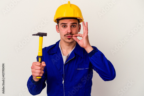 Young caucasian worker man holding a hammer isolated on white background with fingers on lips keeping a secret.