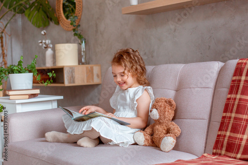 little girl reading a book for teddy bear at home on the couch