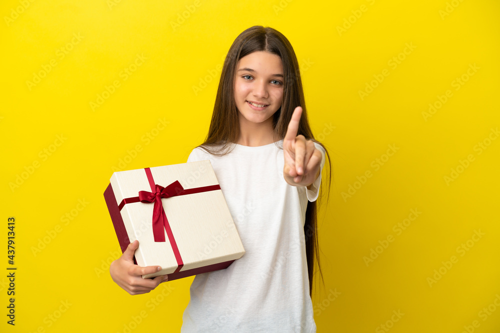 Little girl holding a gift over isolated yellow background showing and lifting a finger Little girl holding a gift over isolated yellow background showing and lifting a finger