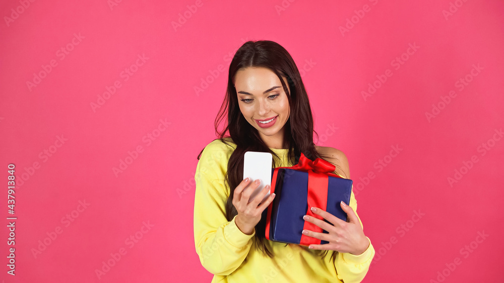 smiling young adult woman holding gift box and looking at cellphone isolated on pink smiling young adult woman holding gift box and looking at cellphone isolated on pink
