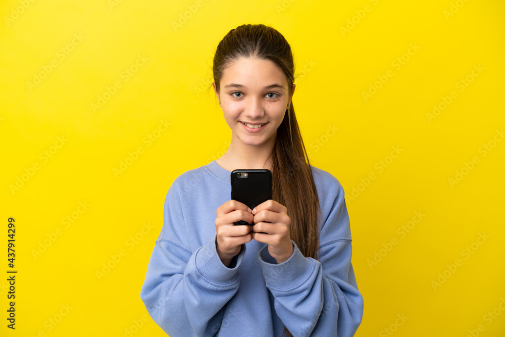 Little girl over isolated yellow background looking at the camera and smiling while using the mobile Little girl over isolated yellow background looking at the camera and smiling while using the mobile