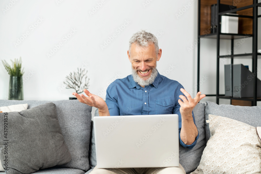 Modern cheerful senior man using laptop for video connection, mature grey haired businessman wearing casual shirt talking, looking into webcam, sitting on the comfortable couch at home Modern cheerful senior man using laptop for video connection, mature grey haired businessman wearing casual shirt talking, looking into webcam, sitting on the comfortable couch at home