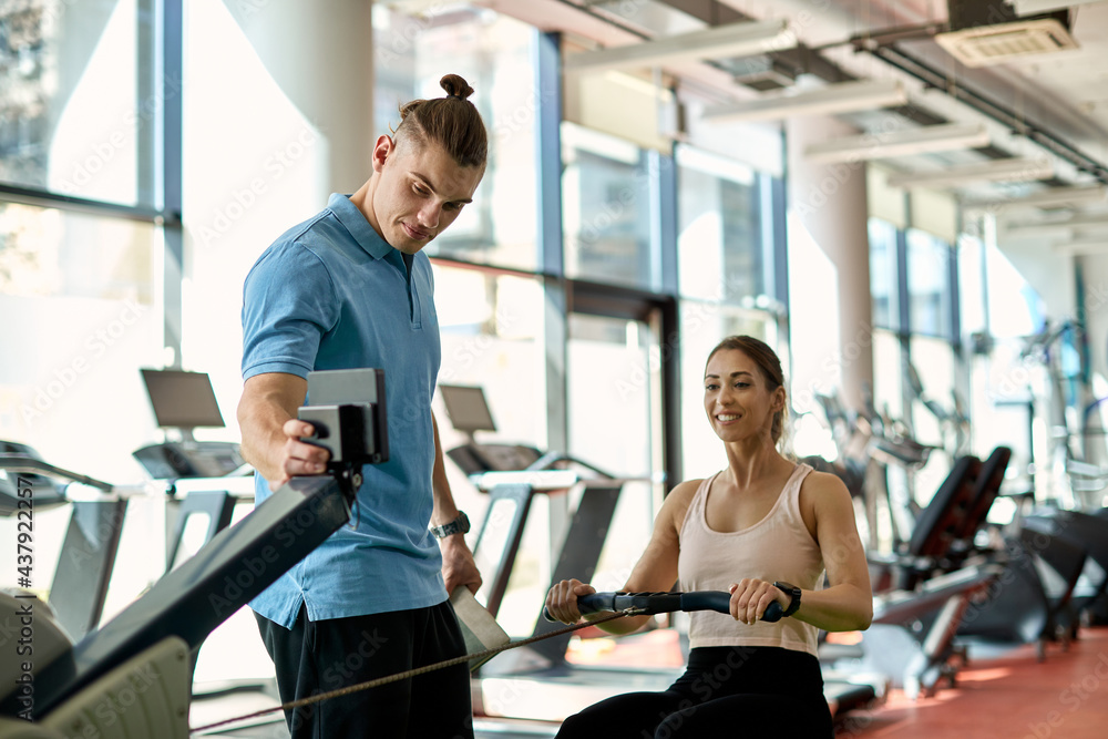 Young athletic woman using rowing machine while having sports training with male coach in a gym.