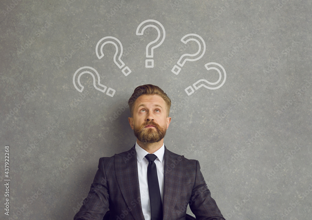 Business man thinking of answers to lots of questions. Confused bearded young man in suit sitting against background of grey concrete wall looking up at several question mark doodles above his head Business man thinking of answers to lots of questions. Confused bearded young man in suit sitting against background of grey concrete wall looking up at several question mark doodles above his head
