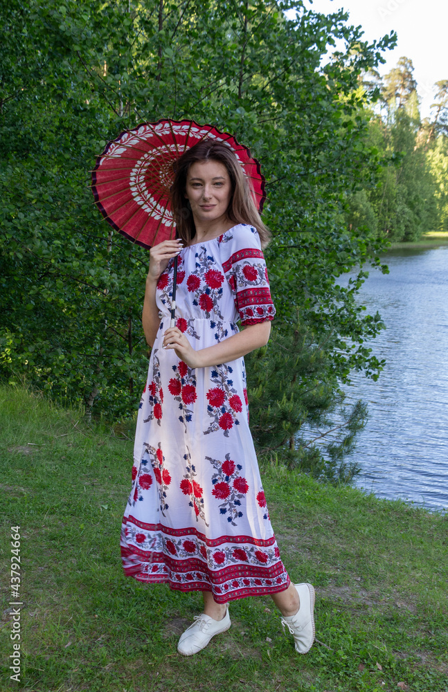 Beautiful young woman in dress with red and white paper umbrella in summer bright day in forest on river side 