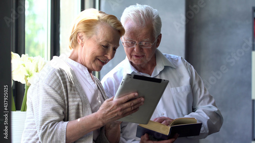 Happy lovely retired couple standing near window using digital tablet