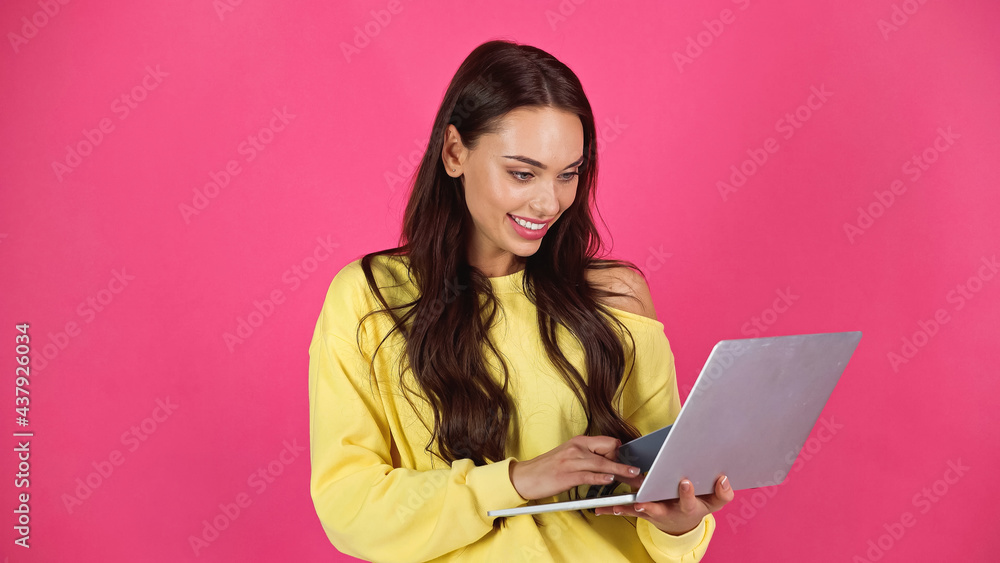Naklejka premium smiling young adult woman typing on laptop and holding credit card isolated on pink