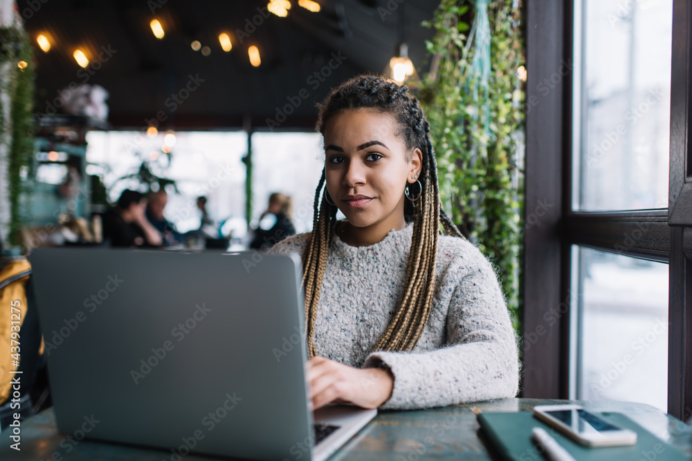 Portrait of dark skinned female millennial sitting at cafeteria table with modern laptop computer and looking at camera, skilled freelance graphic designer using netbook for doing distance job Portrait of dark skinned female millennial sitting at cafeteria table with modern laptop computer and looking at camera, skilled freelance graphic designer using netbook for doing distance job