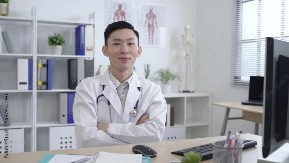 asian physician typing a report at the office desk is pausing to smile at the camera with crossed arms at a bright workplace. asian physician typing a report at the office desk is pausing to smile at the camera with crossed arms at a bright workplace.